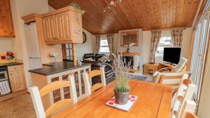 Un acogedor interior de cabaña con una mesa de comedor de madera, sillas, una zona de cocina, una sala de estar con chimenea y un televisor.
