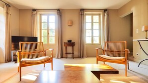 Una sala de estar acogedora con dos sillones de madera, una mesa de centro de madera, un televisor y ventanas amplias con cortinas.
