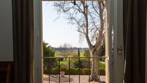 Vista desde una habitación a través de una puerta abierta, mostrando una barandilla metálica, un árbol grande y un jardín cuidado con una planta en maceta.