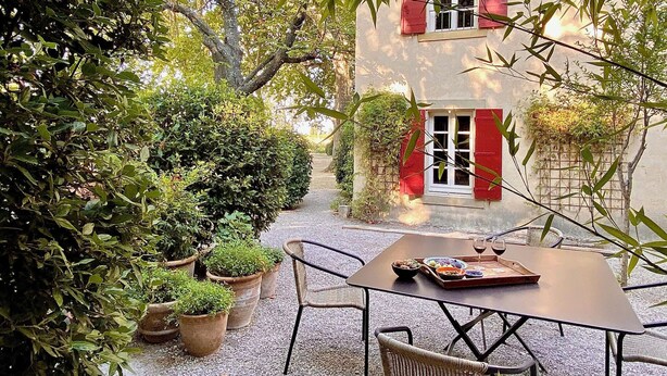 Un espacio al aire libre con mesa y sillas, rodeado de plantas en macetas y un edificio con contraventanas rojas.