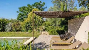 Un área de piscina con sillones de descanso bajo una pérgola, rodeada de follaje.