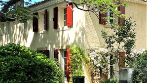 Un edificio tradicional con contraventanas rojas, un jardín con diversas plantas y un muro de piedra.