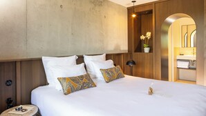 A hotel room with a large bed, wooden headboard, a small table with a lamp, and a bathroom visible through an archway. - Son de Mar (Lecci)