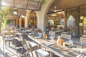 Un área de comedor al aire libre con mesas y sillas, un ventilador de techo y vistas del interior a través de puertas de vidrio.
