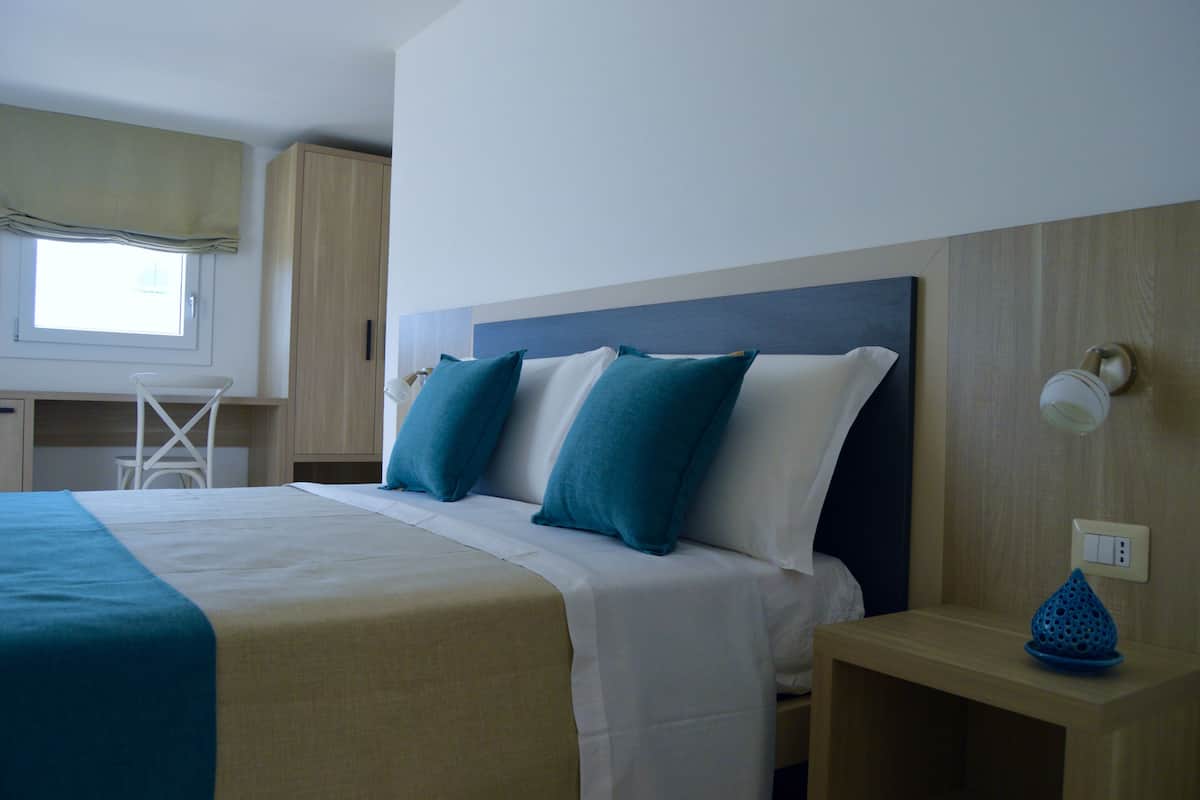 Uma cama bem arrumada com esquema de cores azul e branco, uma mesinha de cabeceira pequena com um objeto decorativo azul e uma janela com um valance verde.