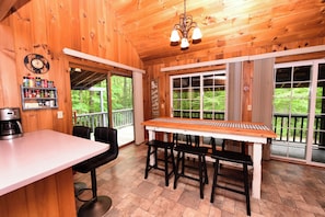 A wooden interior with a kitchen area, bar stools, a coffee maker, and large windows with a view of greenery.