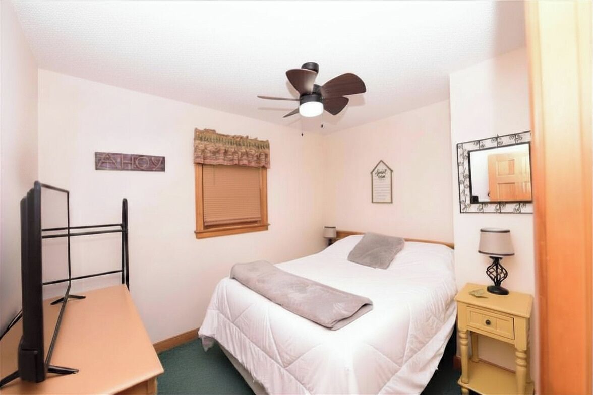 A bedroom with a bed, a desk, a chair, a ceiling fan, a window with blinds, a bedside table with a lamp, and a mirror on the wall.