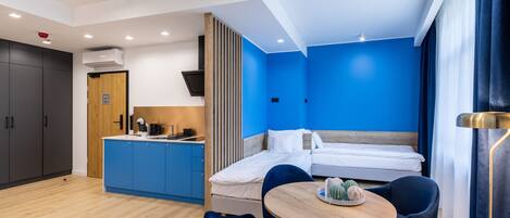 A modern hotel room with a blue accent wall, a wooden dining table, and a separate bed area.