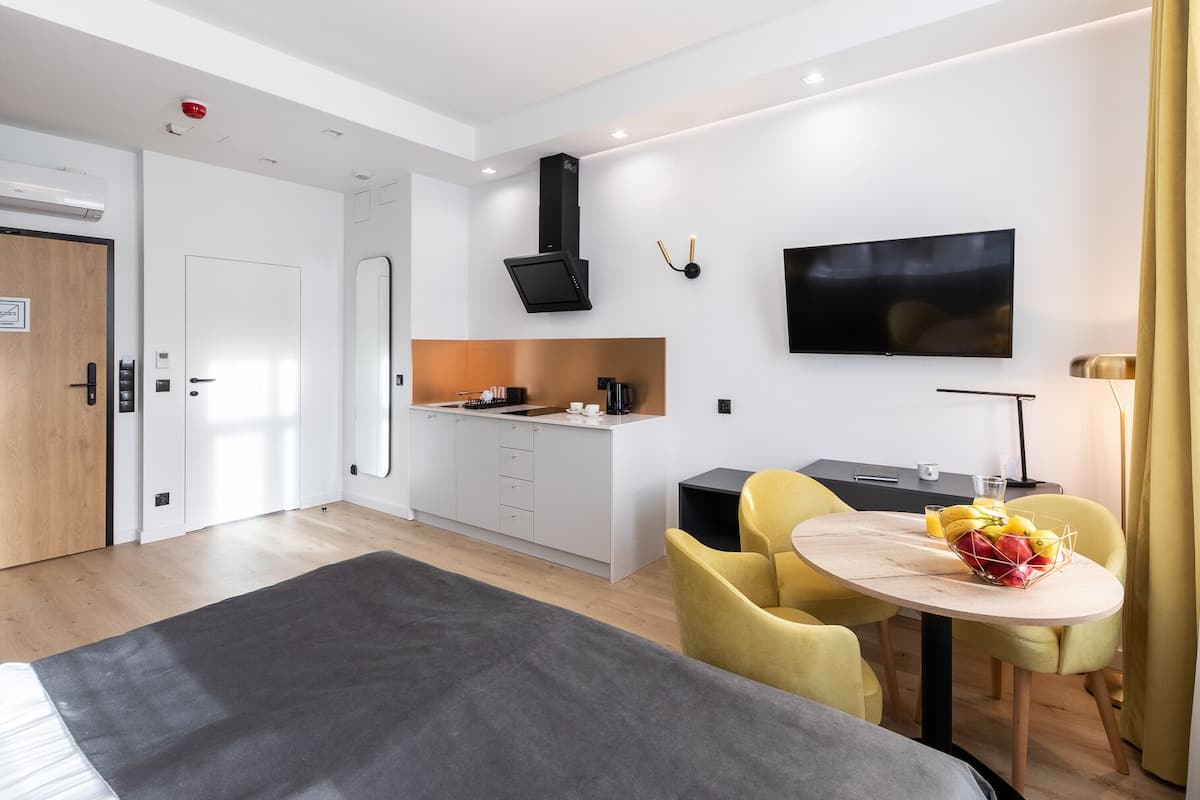Una habitación de hotel moderna con una cama, una zona de comedor con sillas amarillas, un televisor y una kitchenette.