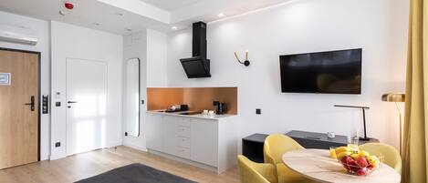A modern hotel room with a bed, a dining area with yellow chairs, a TV, and a kitchenette.