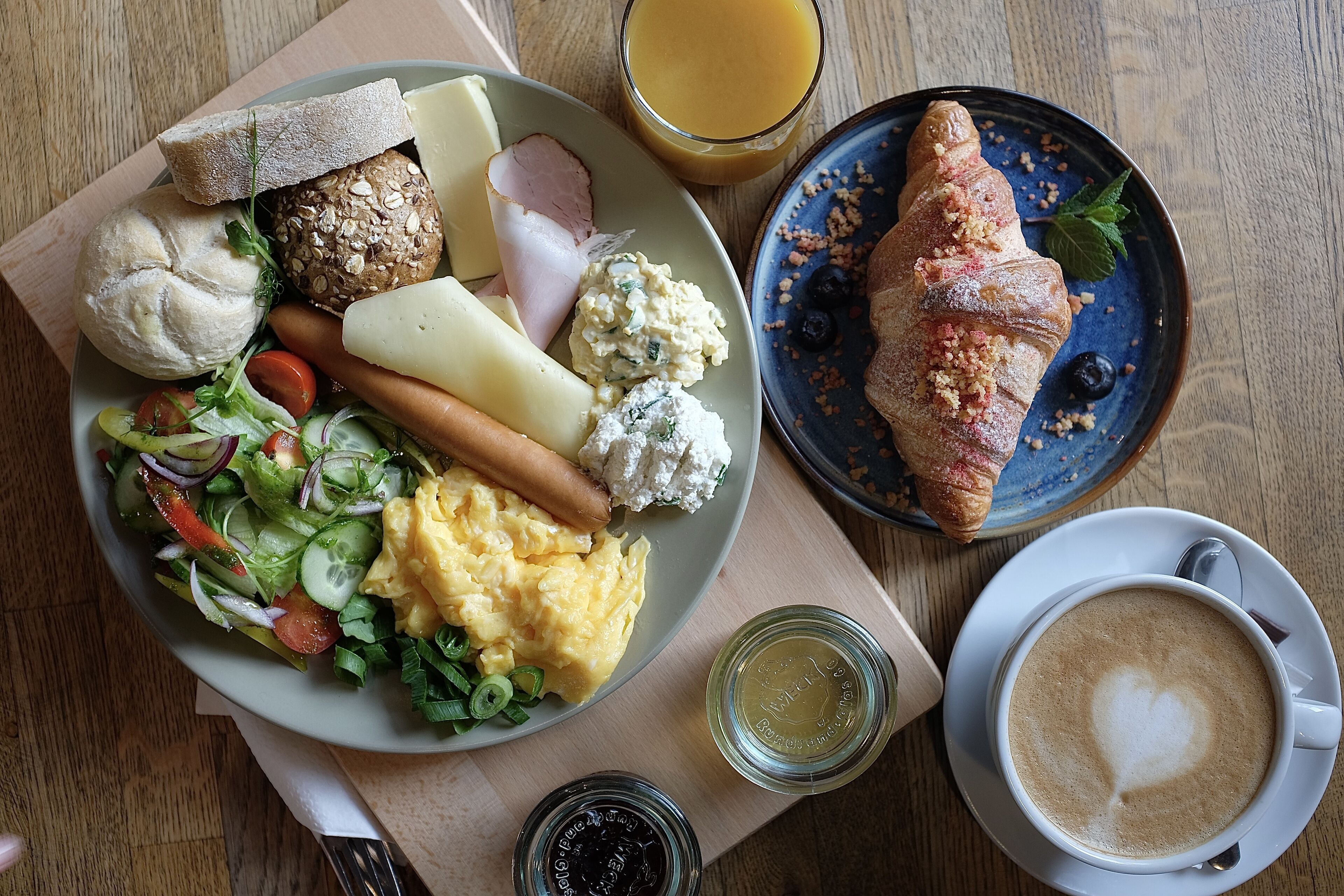 Un plato de desayuno con huevos, jamón, pan y un croissant, junto con una taza de café y un vaso de jugo de naranja.