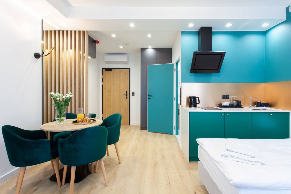 Habitación de hotel moderna con cama, zona de comedor con mesa y sillas, cocina con fregadero y estufa, y un televisor sobre el mostrador.