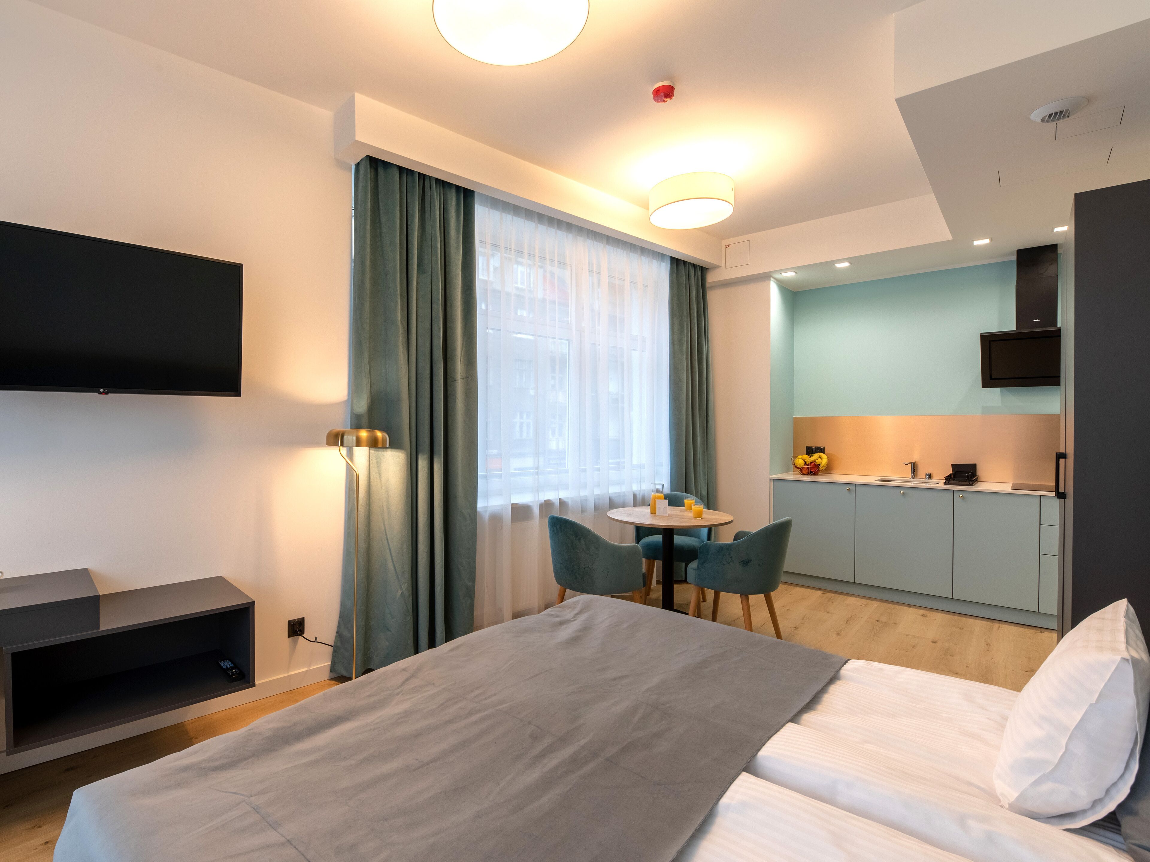 Ein modernes Hotelzimmer mit Bett, Schreibtisch, Stuhl, Flachbildfernseher, einer kleinen Küchenzeile und einem fenster mit Vorhängen.
