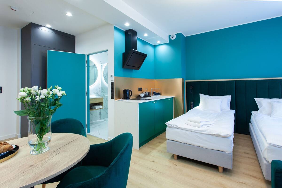 Una habitación de hotel moderna con una cama, una kitchenette pequeña, una mesa de comedor con sillas y un jarrón de flores.