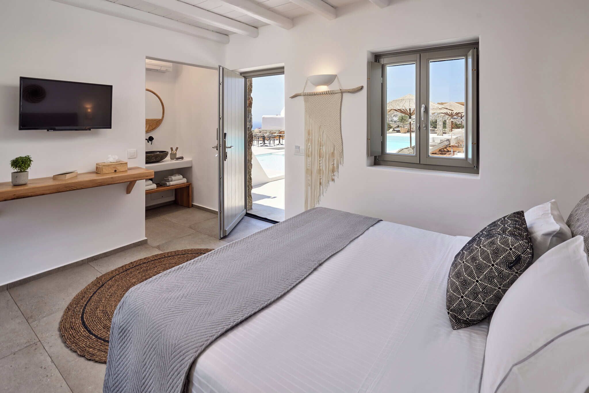 Un dormitorio moderno con cama, televisor, estante de madera, macramé colgante y vista a una piscina al aire libre.