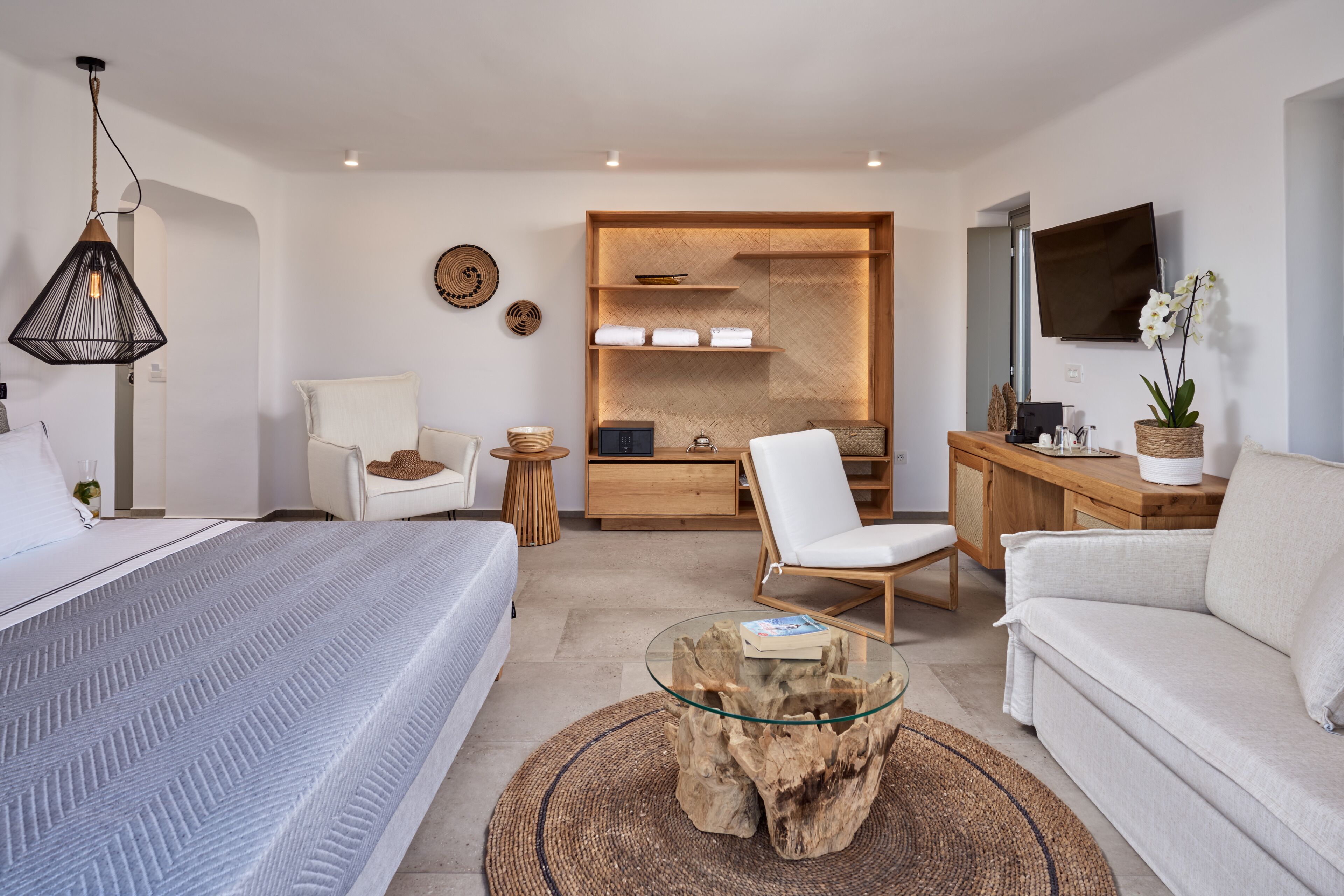 A modern hotel room with a bed, a glass coffee table, a white sofa, a wooden desk, and a TV mounted on the wall.