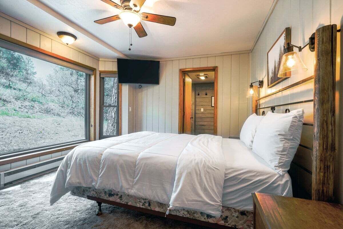 A bedroom with a bed, a ceiling fan, a TV, and a large window.