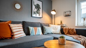 Una sala de estar moderna con un sofá gris, varios cojines, una mesa redonda de madera con una planta en maceta, una lámpara de pie y una obra de arte con motivos vegetales en la pared.