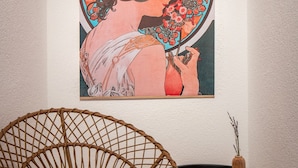 Una habitación con una silla de mimbre, una mesita con un jarrón y una obra de arte grande enmarcada en la pared.