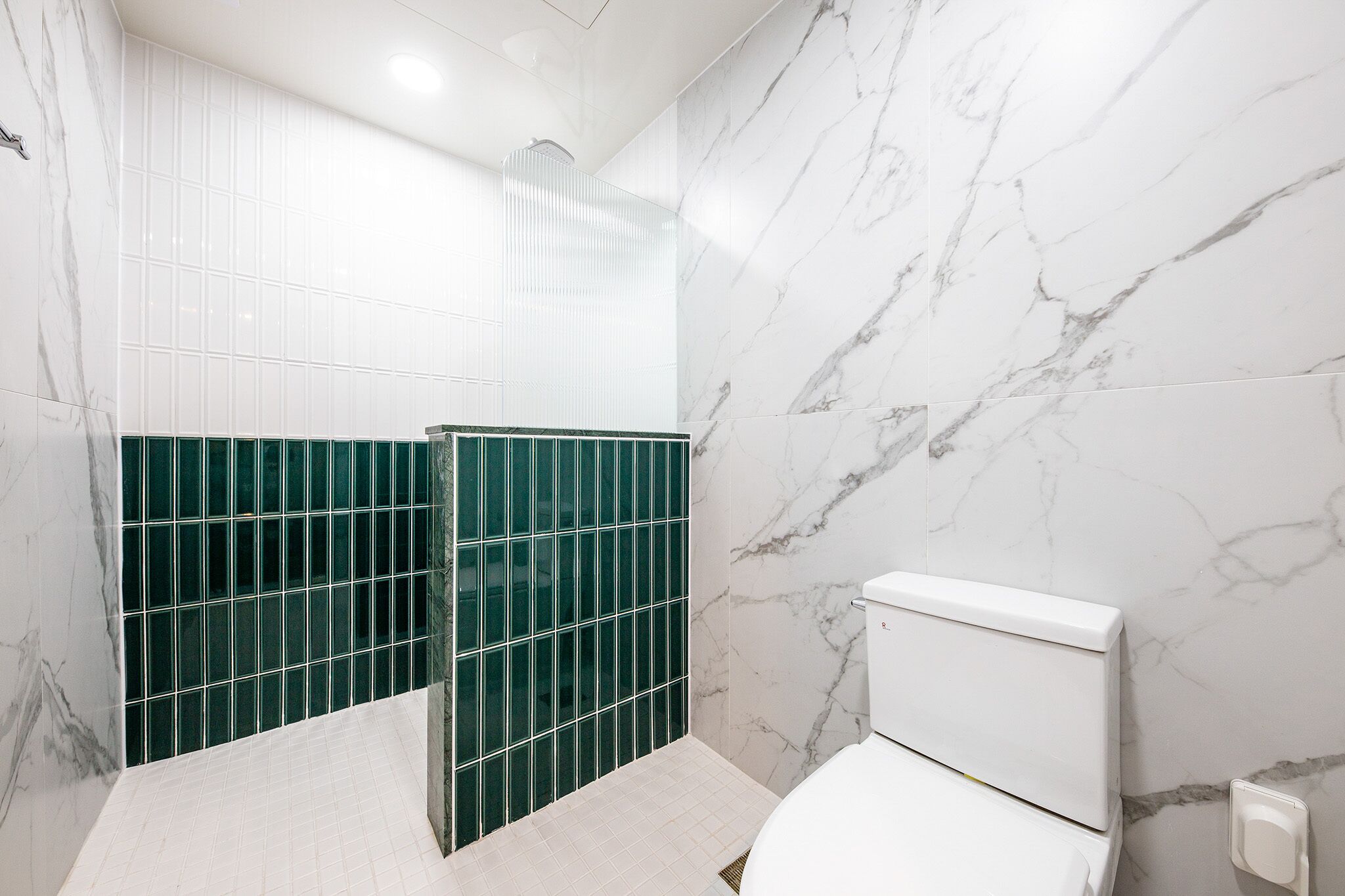 Un baño con paredes de mármol blanco, cabina de ducha con azulejos verdes y un inodoro blanco.