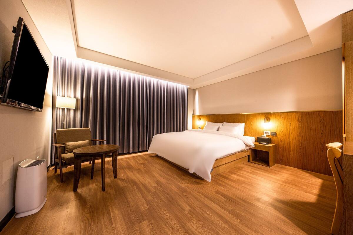 Une chambre d’hôtel avec un lit, une chaise, une table, un téléviseur, une lampe et un plancher en bois.