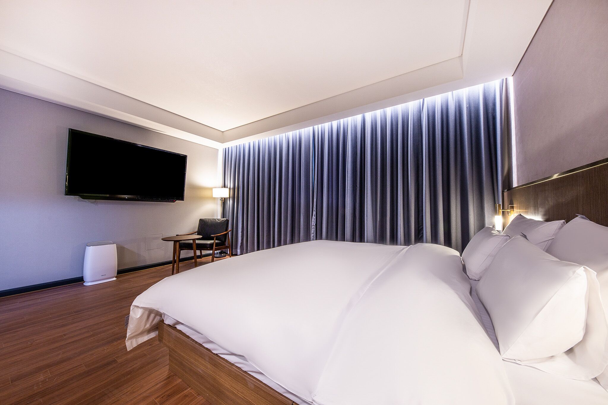 Ein modernes Hotelzimmer mit einem großen Bett, einem flachbildfernseher an der Wand, einem Schreibtisch mit Stuhl und einem großen Fenster mit Vorhängen.