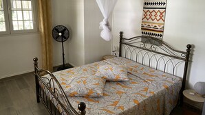 Ein Schlafzimmer mit einem Bett, einem Standventilator, einem Moskitonetz, einem Wandbehang mit Muster sowie einem Fenster mit Vorhängen.