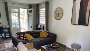 Ein Wohnzimmer mit einer Couch, einem Couchtisch, einem Sessel und einem kleinen Hocker. Eine Tür führt zu einem Außenbereich.
