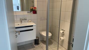 Un baño moderno con cabina de ducha de vidrio, un lavabo blanco con espejo y un inodoro con dispensador de toallas de papel.