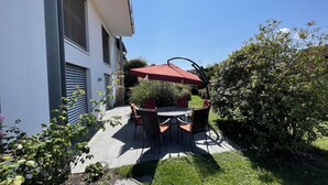 Un patio con mesa y sillas, una sombrilla roja y un jardín con diversas plantas.