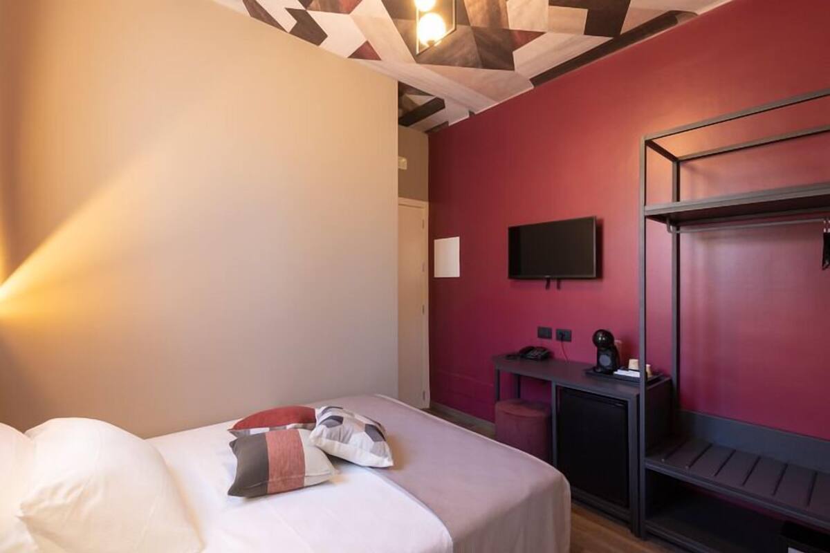 A hotel room with a bed, a desk with a chair, a TV mounted on the wall, and a red accent wall.