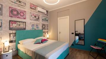 A bedroom with a teal headboard, a bed with white and teal bedding, a wall decorated with vintage cassette tapes, a mirror, and a rainbow-striped chair.