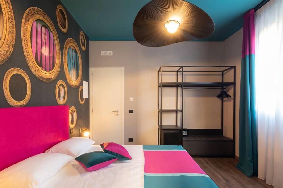 A hotel room with a bunk bed, a bed with colorful bedding, a wall with framed mirrors, and a ceiling light fixture.