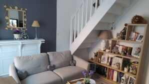 Un salon avec un canapé beige, une petite table blanche sur laquelle se trouve un plateau, une bibliothèque et un escalier.