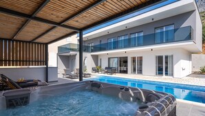 A modern outdoor pool area with a hot tub, lounge chairs, and a covered patio.