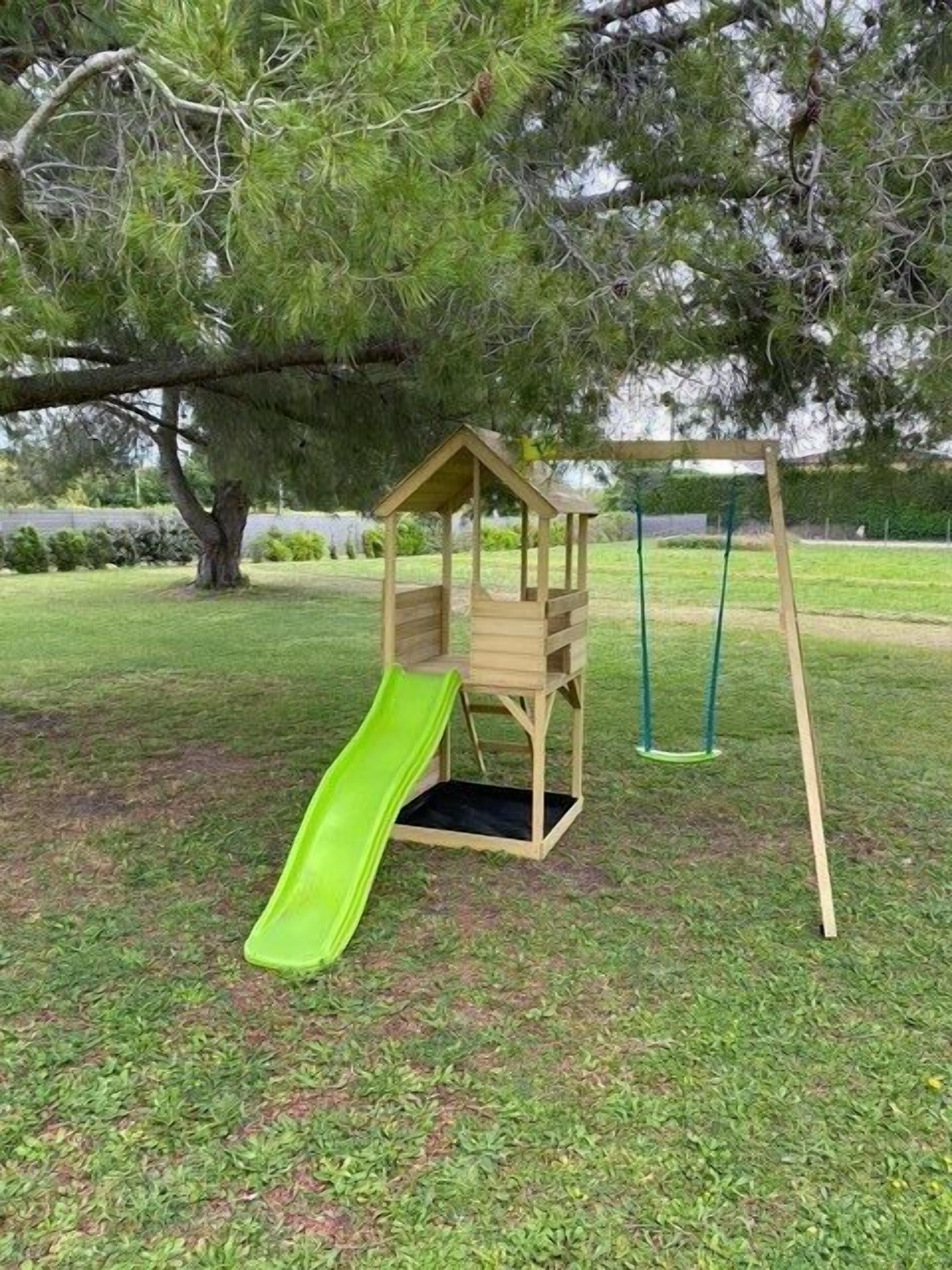 Una casita de juegos de madera con un tobogán verde en un parque cubierto de césped.