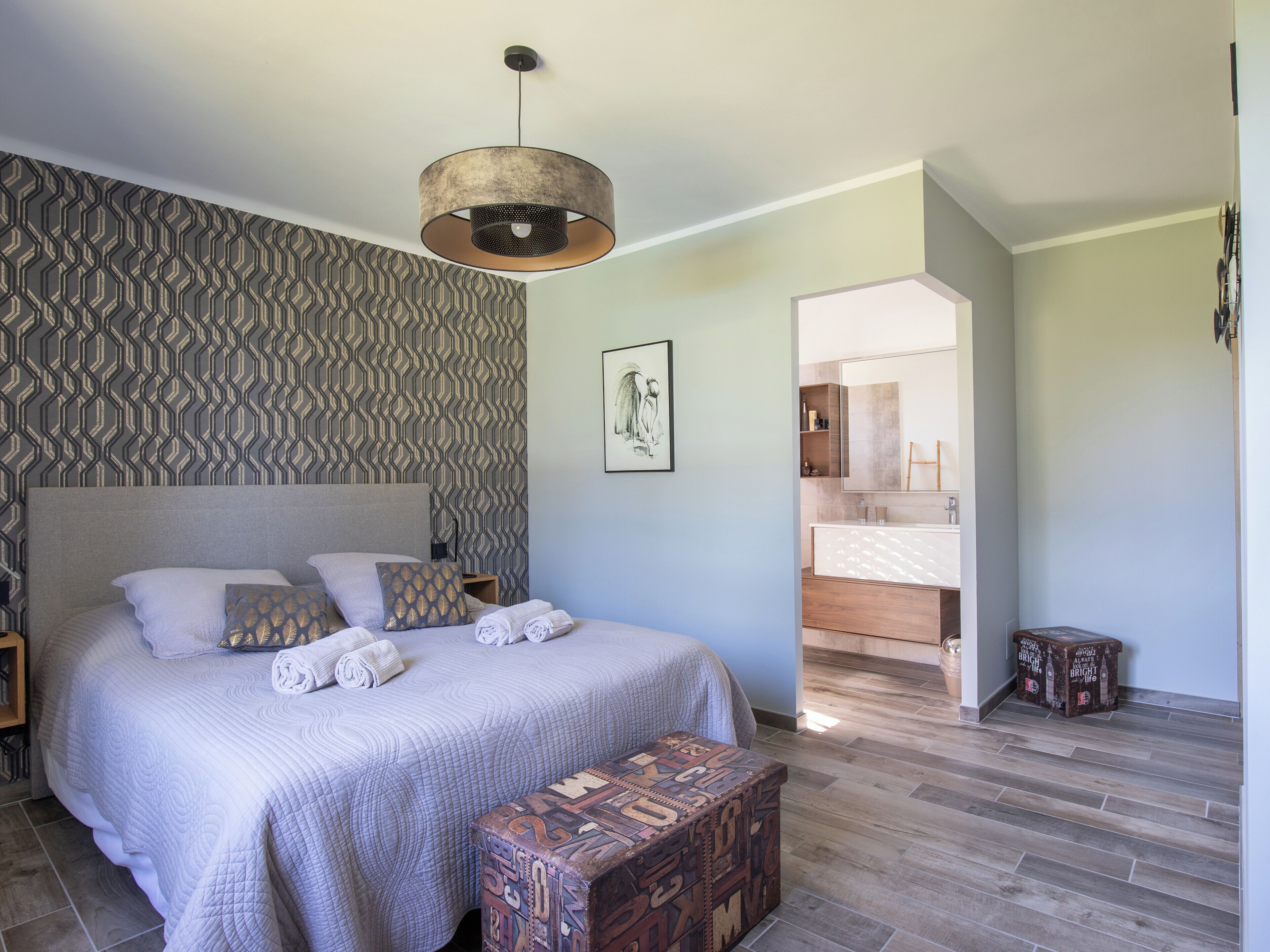 A bedroom with a large bed, patterned wallpaper, a hanging light fixture, and a wooden bench.