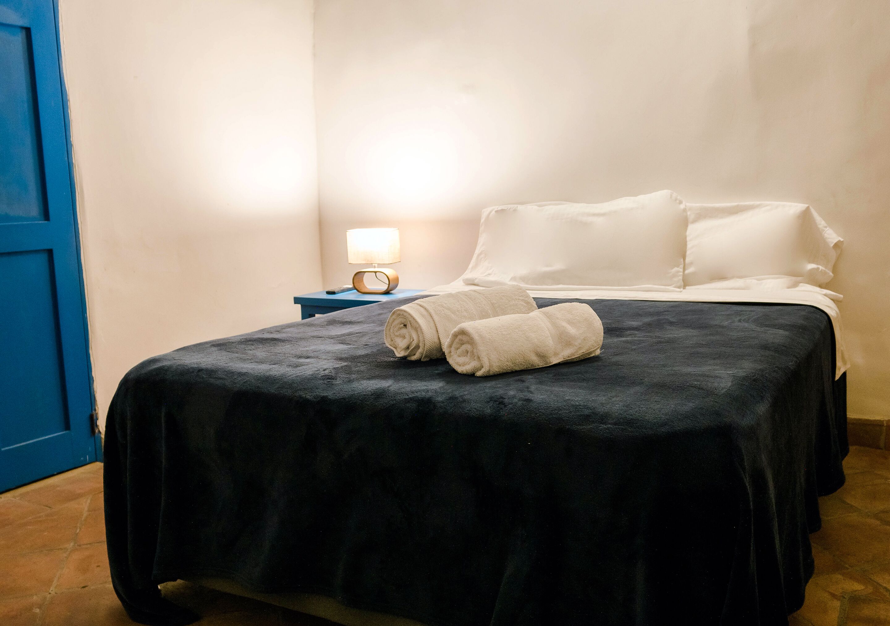 A neatly made bed with a black bedspread and white pillows, a bedside lamp, and a blue door.