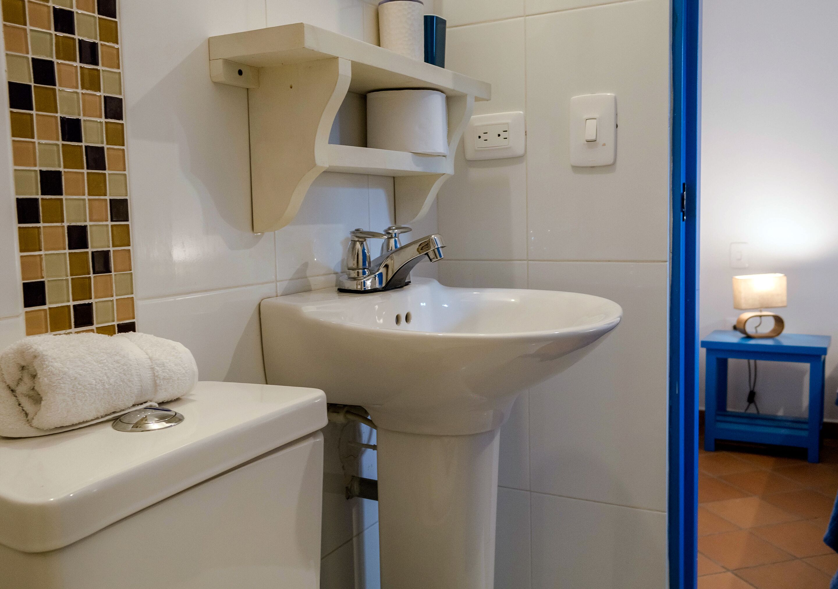 A bathroom with a white sink, toilet, and a towel on the toilet. There is a shelf above the sink holding a toilet paper roll and a small container.