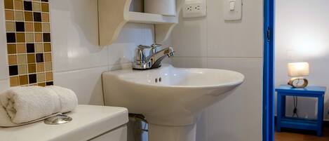 A bathroom with a white sink, toilet, and a towel on the toilet. There is a shelf above the sink holding a toilet paper roll and a small container.