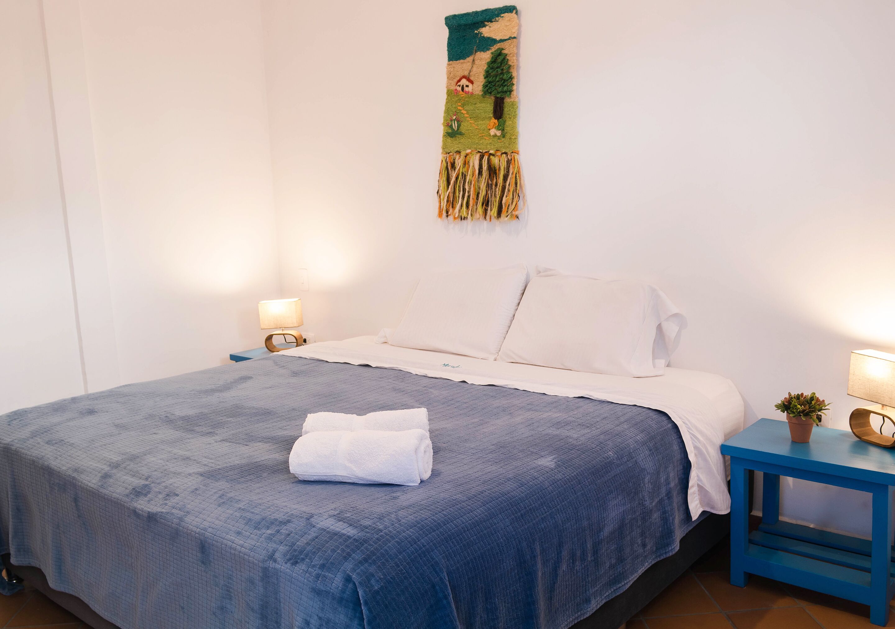 Un lit bien fait, avec une couvre-lit bleue et des oreillers blancs, une petite table de chevet avec une lampe, et une décoration murale au-dessus du lit.