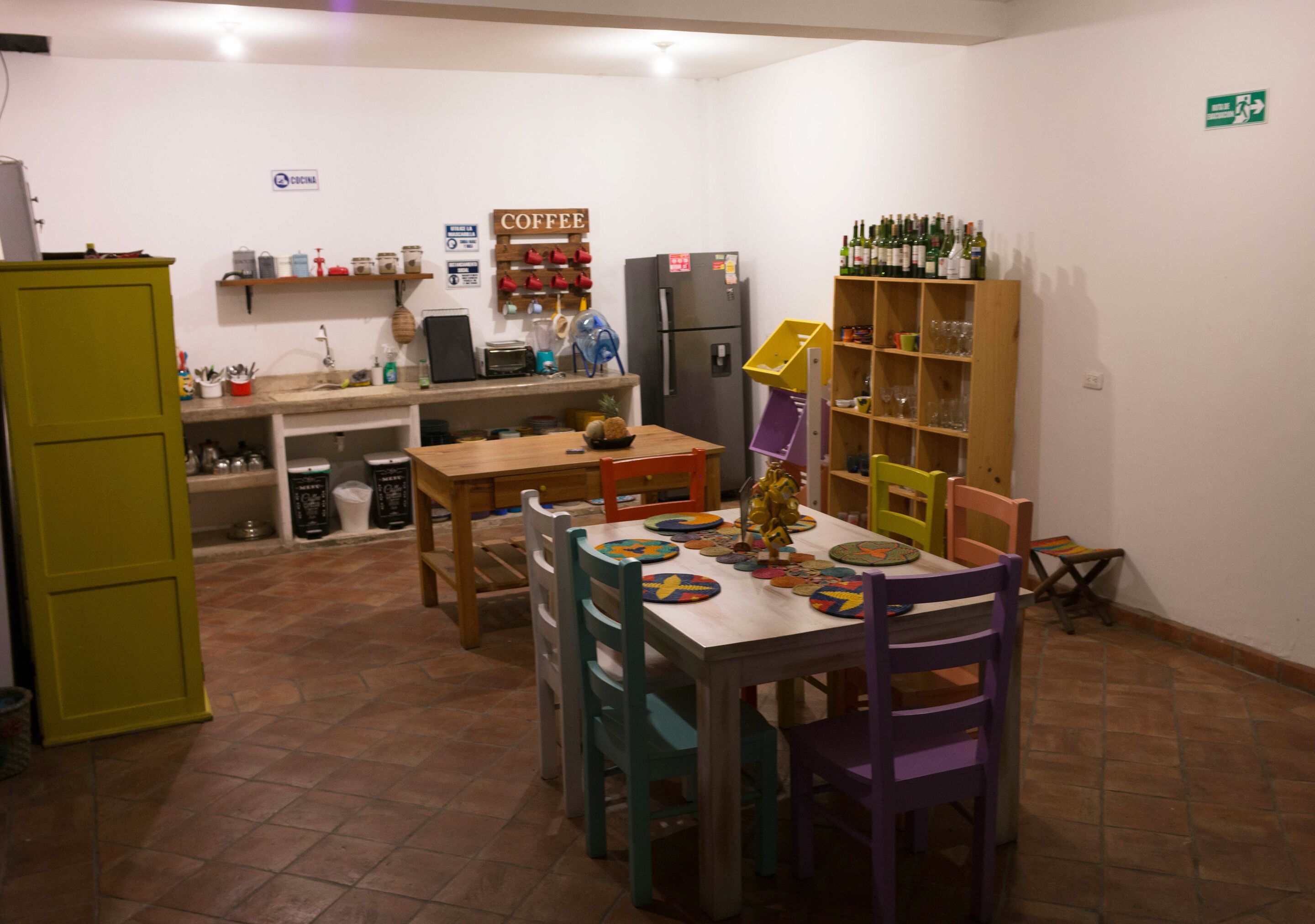 Cocina compacta con comedor, equipada con refrigerador, estación de café y repisas con botellas y vasos.