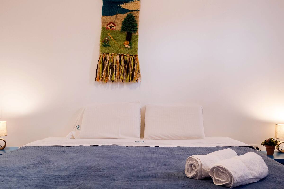 Un llit ben fet amb un llençol blau i coixins blancs, una tovallola enrotllada sobre el llit, i un tapís de paret amb una escena de paisatge.