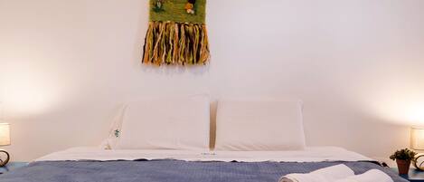 A neatly made bed with a blue bedspread and white pillows, a rolled towel on the bed, and a wall tapestry depicting a landscape scene.