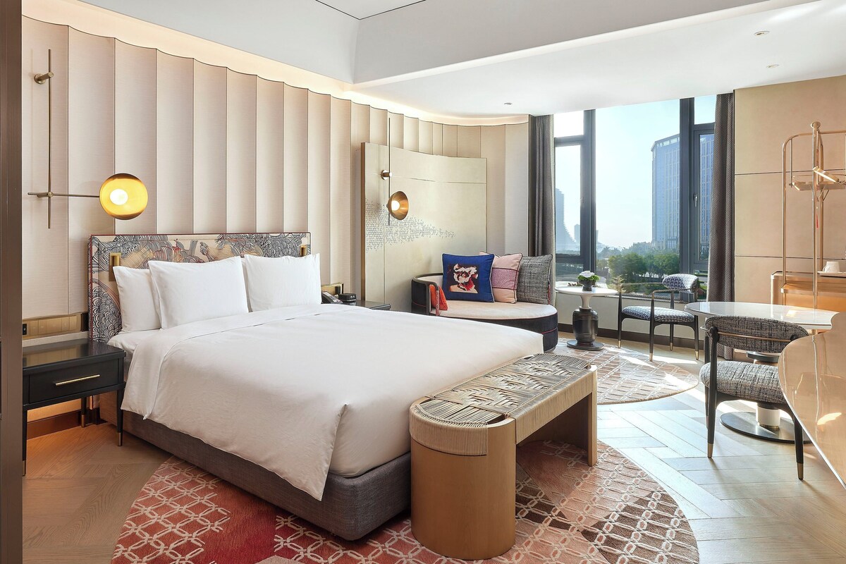 A modern hotel room with a large bed, a sitting area with a table and chairs, and a view of the city through large windows.