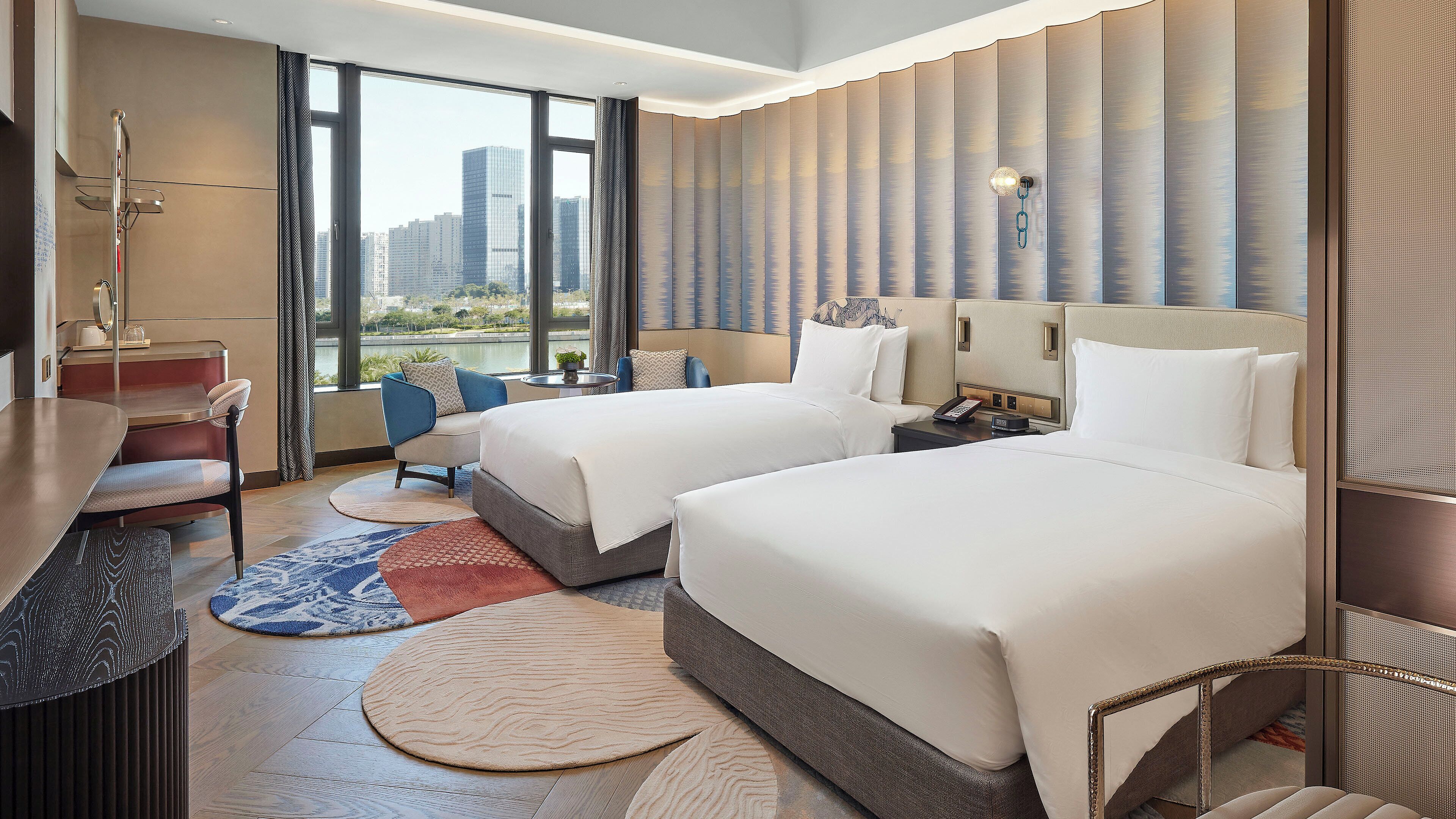 Habitación de hotel con dos camas, un escritorio, una silla y vista a un paisaje urbano.