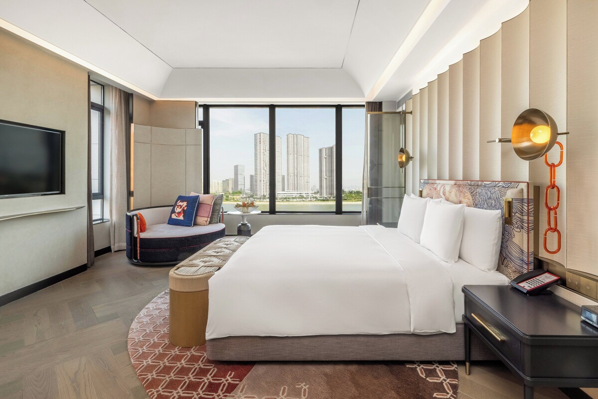 A modern hotel room with a large bed, a seating area, a TV, and a city view.