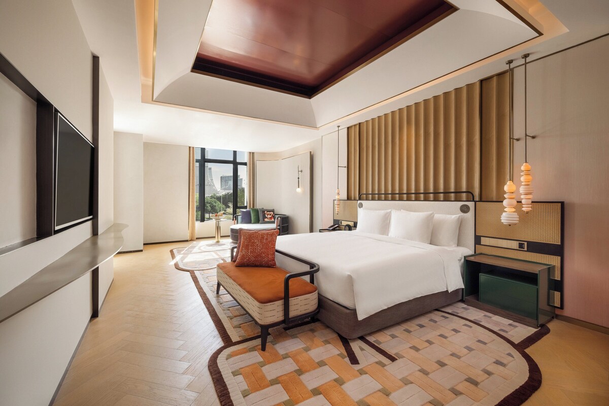 A modern hotel room with a large bed, a TV, a desk, and a view of the city.
