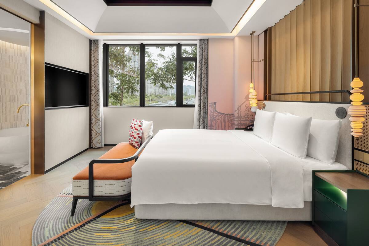 A modern hotel room with a large bed, a TV, a seating area, and a view of the city.
