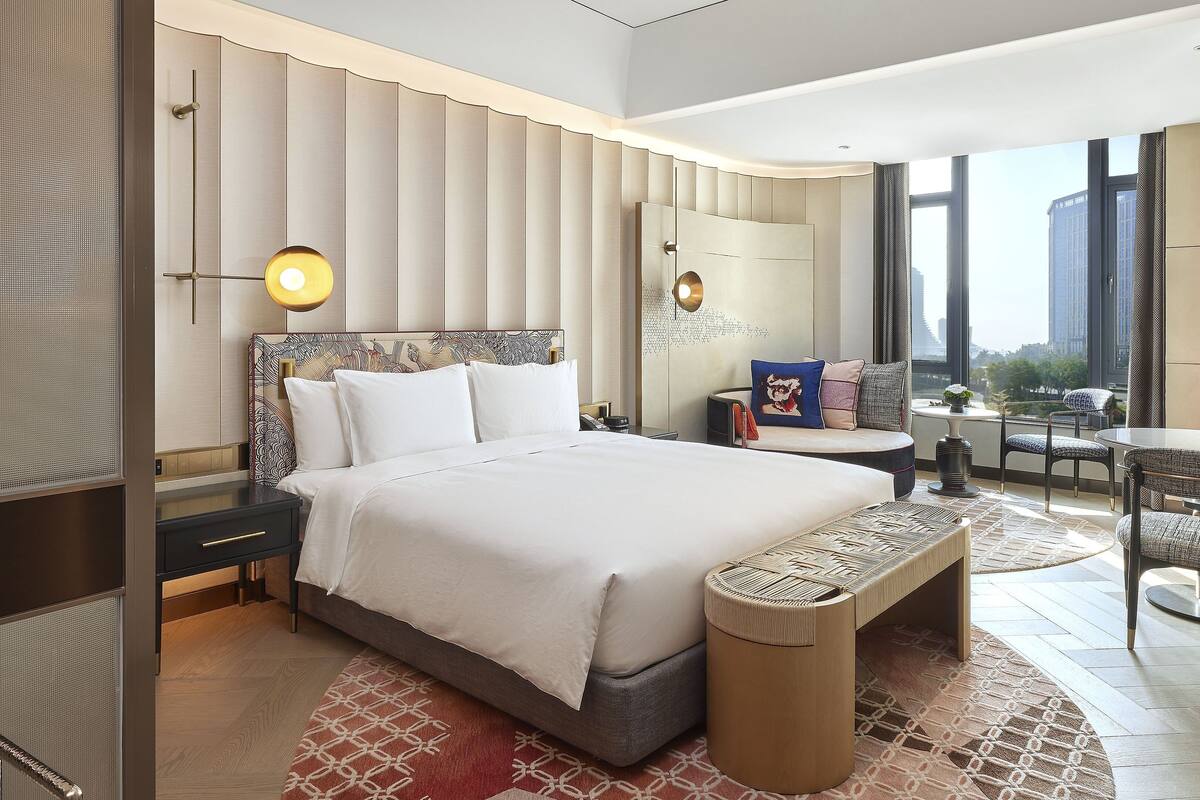 A modern hotel room with a large bed, a sitting area with a table and chairs, and a view of the city through large windows.
